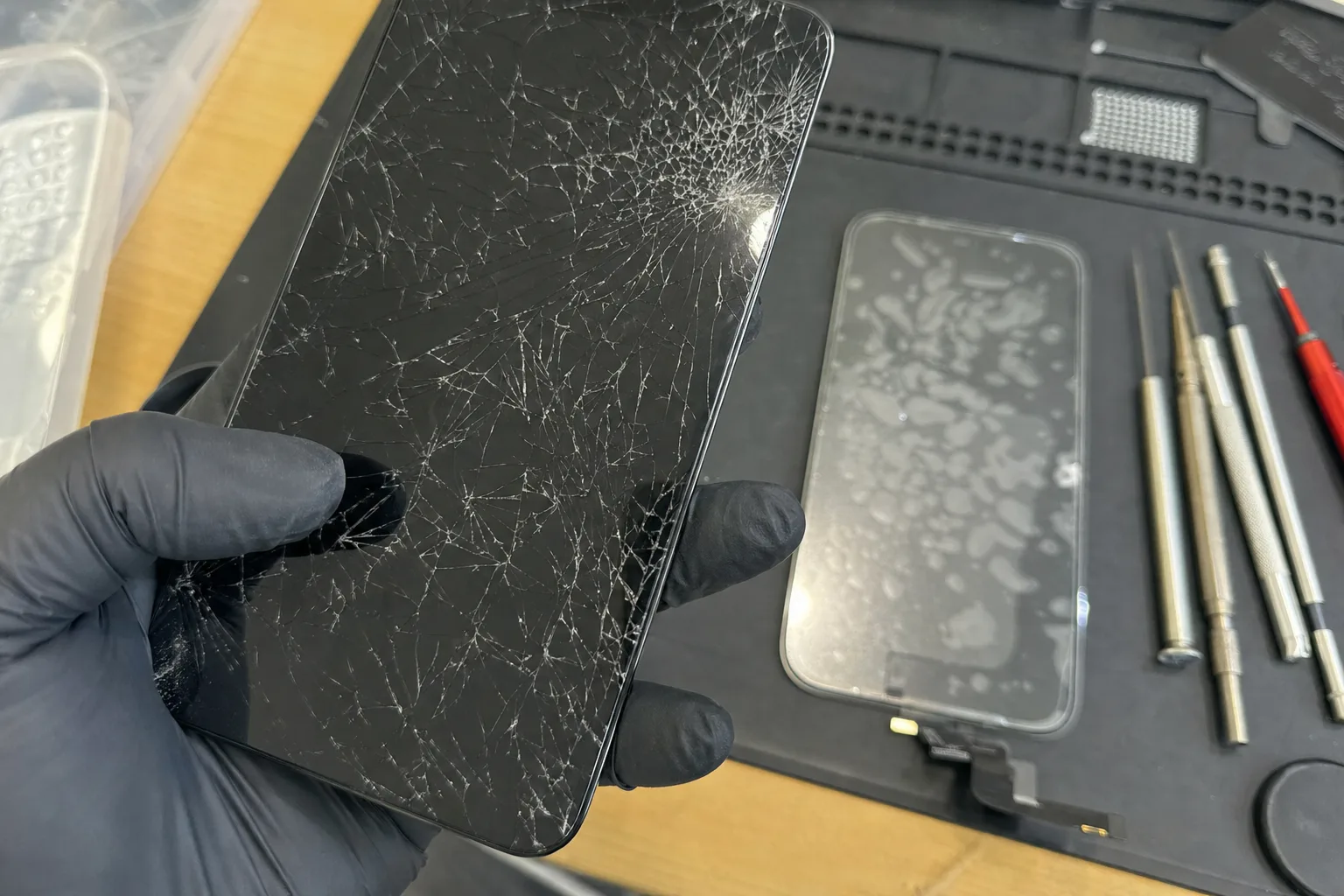 iPhone à l'écran fissuré tenu au-dessus d'un plan de travail d'atelier, écran neuf prêt à poser